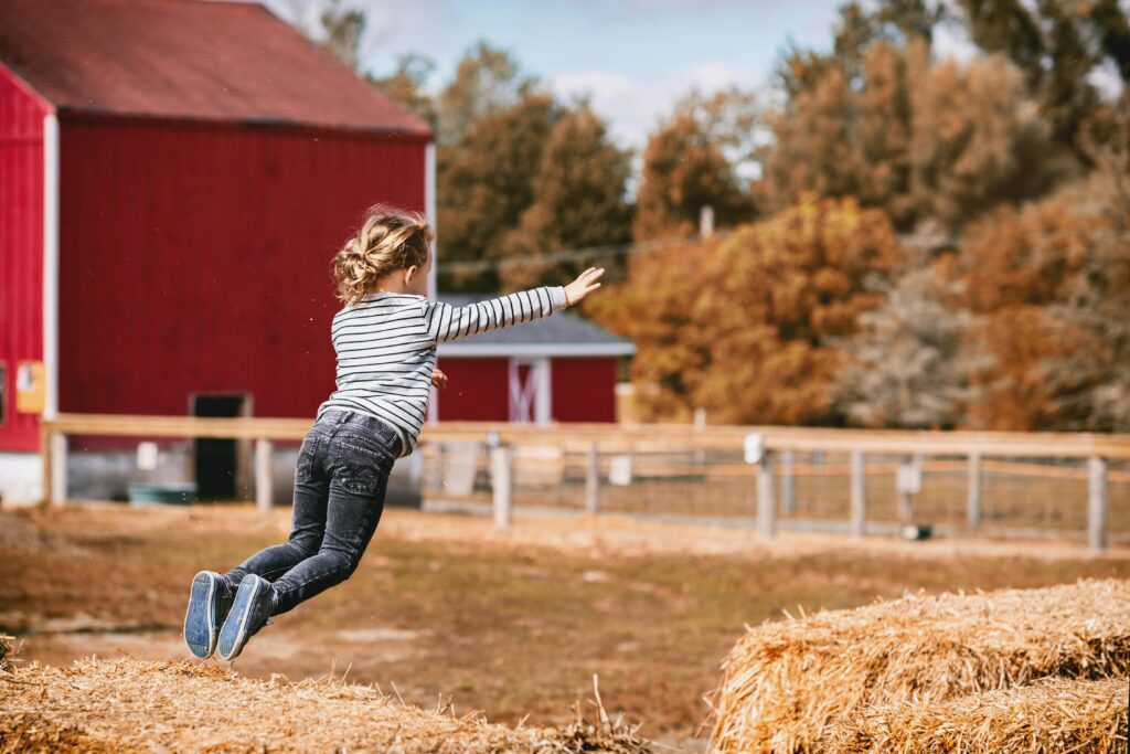 Young girl joyfully jumping on hay bales in a rural countryside setting during fall.