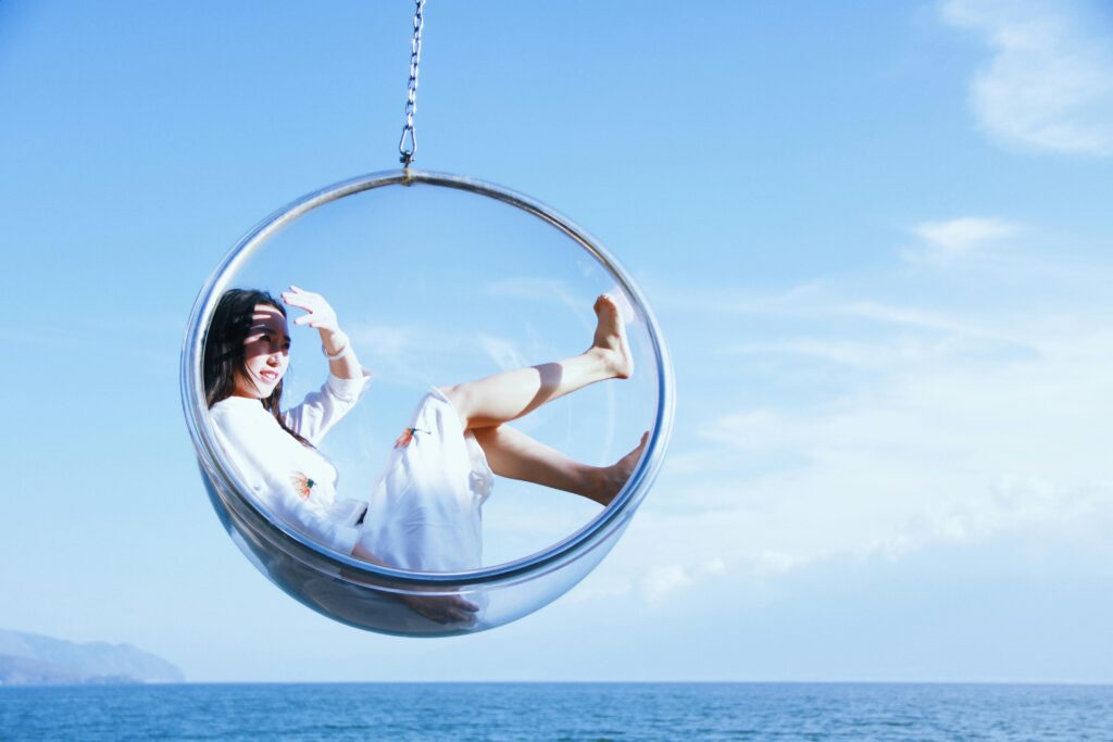 A woman enjoys a sunny day in a hanging bubble chair by the ocean, perfect for a relaxing vacation.