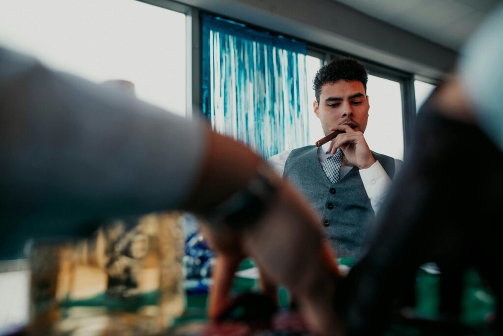 A stylish man holds a cigar while seated at an indoor gathering, exuding a sophisticated ambiance.